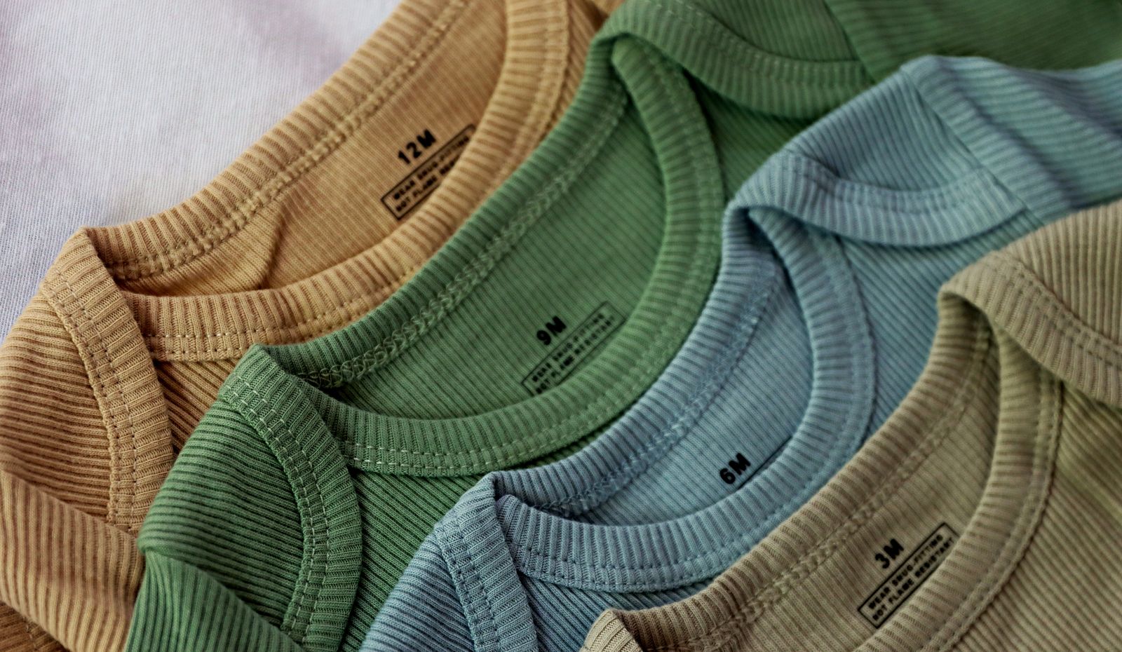 Four ribbed bodysuits fanned out in ochre, green, blue and oat, each with its size tag visible.