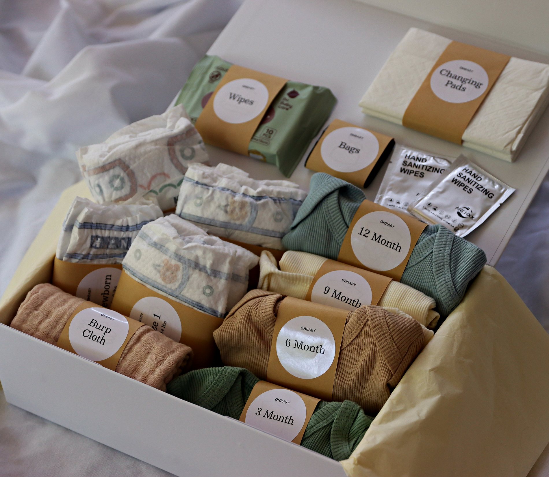The Oh Baby Kit gift box open on soft bedding, packed with diapers labeled Newborn through Size 3, ribbed bodysuits labeled 3 to 12 months, wipes, changing pads, burp cloth and diaper bags.