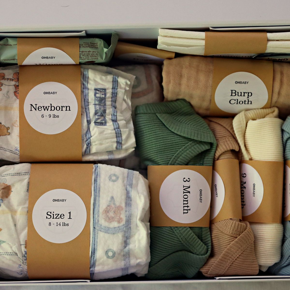 Overhead view of the packed Oh Baby Kit: diaper bundles labeled Newborn 6-9 lbs and Size 1 8-14 lbs, a burp cloth, and ribbed bodysuits labeled by month, each banded with a kraft label.