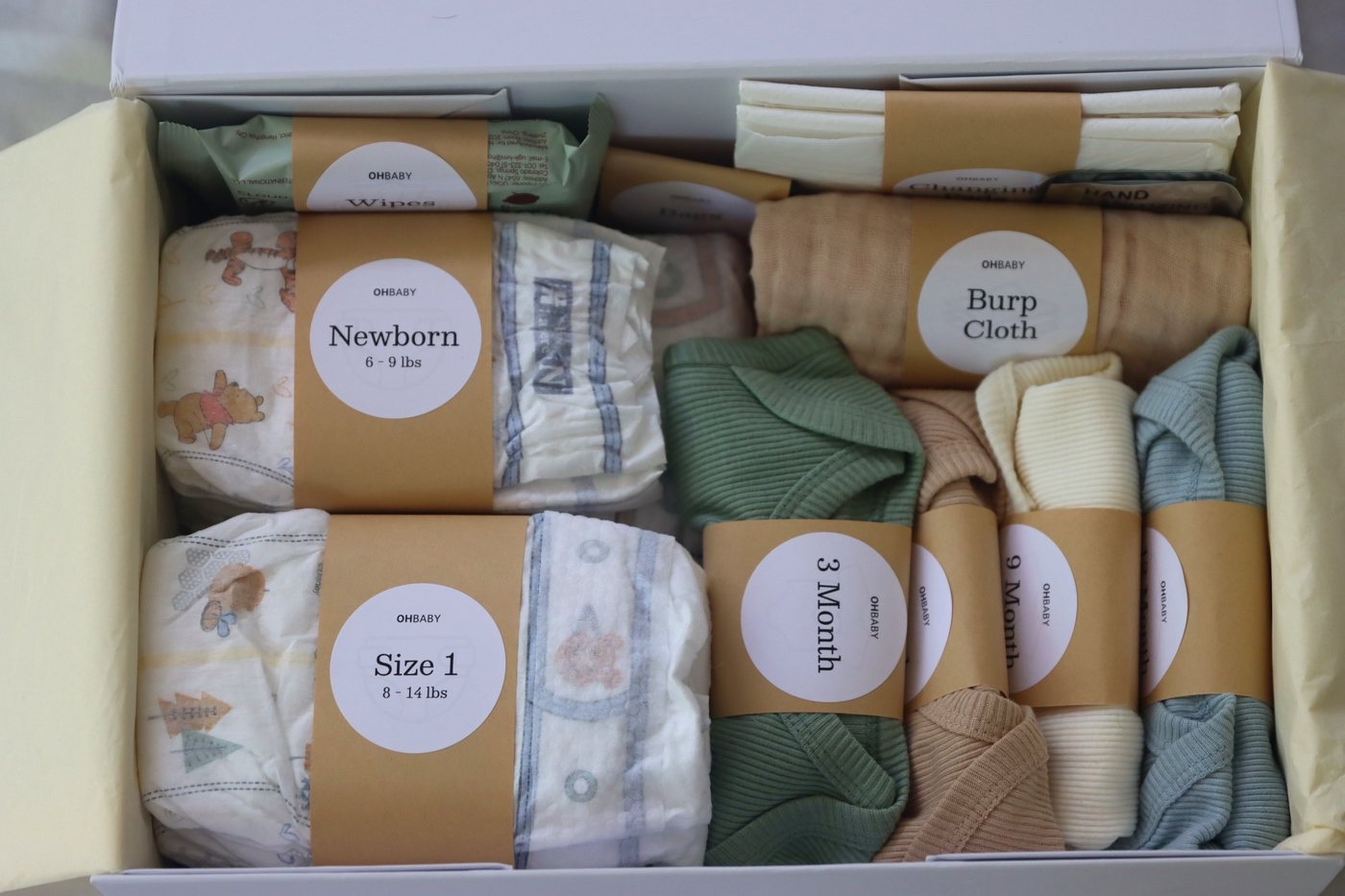 Inside the Oh Baby Kit box: diapers and bodysuits wrapped in kraft bands, each labeled by size and month.